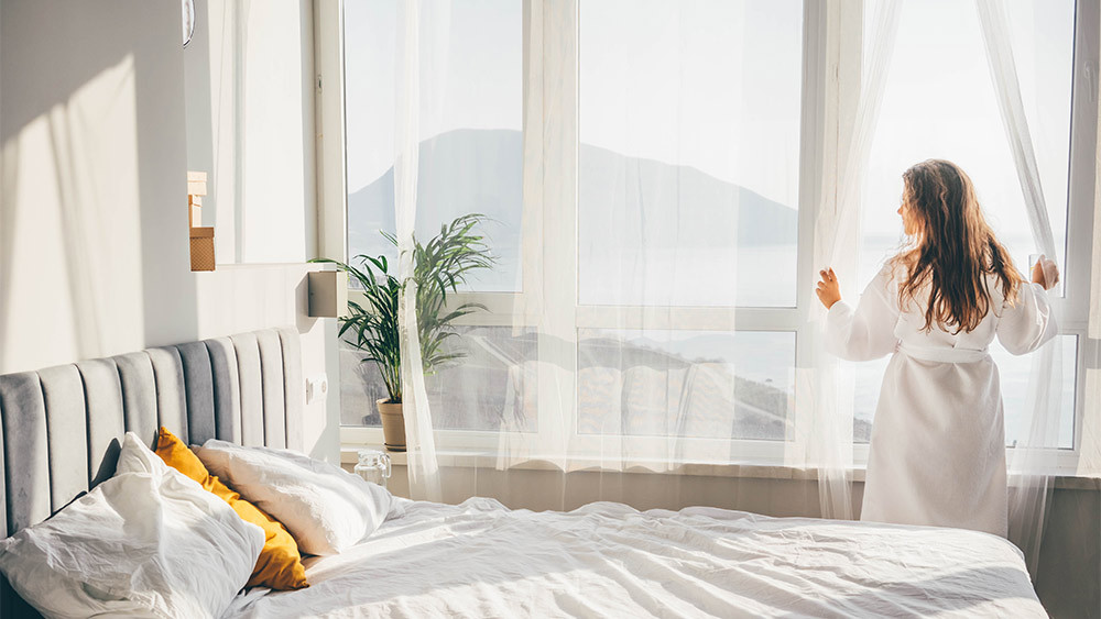 A woman opening blinds to let the sun in and to help warm up a room without a heater.