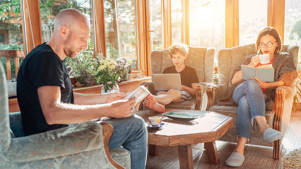 A man and a woman are reading while a kid is working on his computer in a sunroom