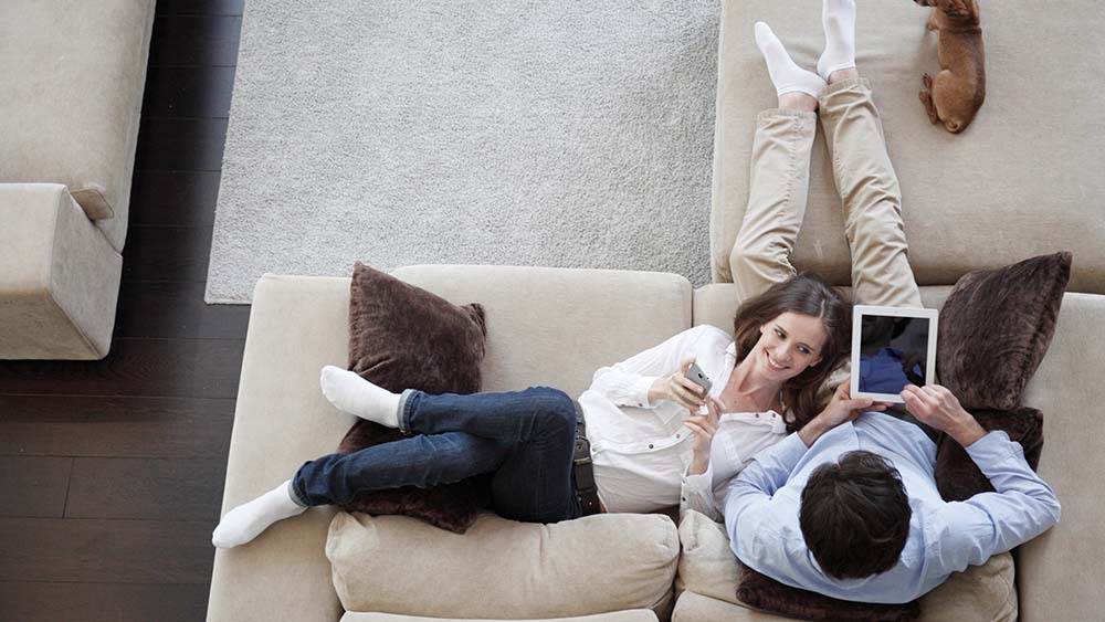 A man is using his tablet while lying on a couch while a woman is relaxing nearby