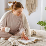 woman sitting on bed, sipping tea and reading a book