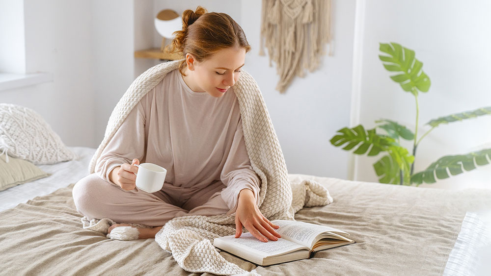 woman sitting on bed, sipping tea and reading a book